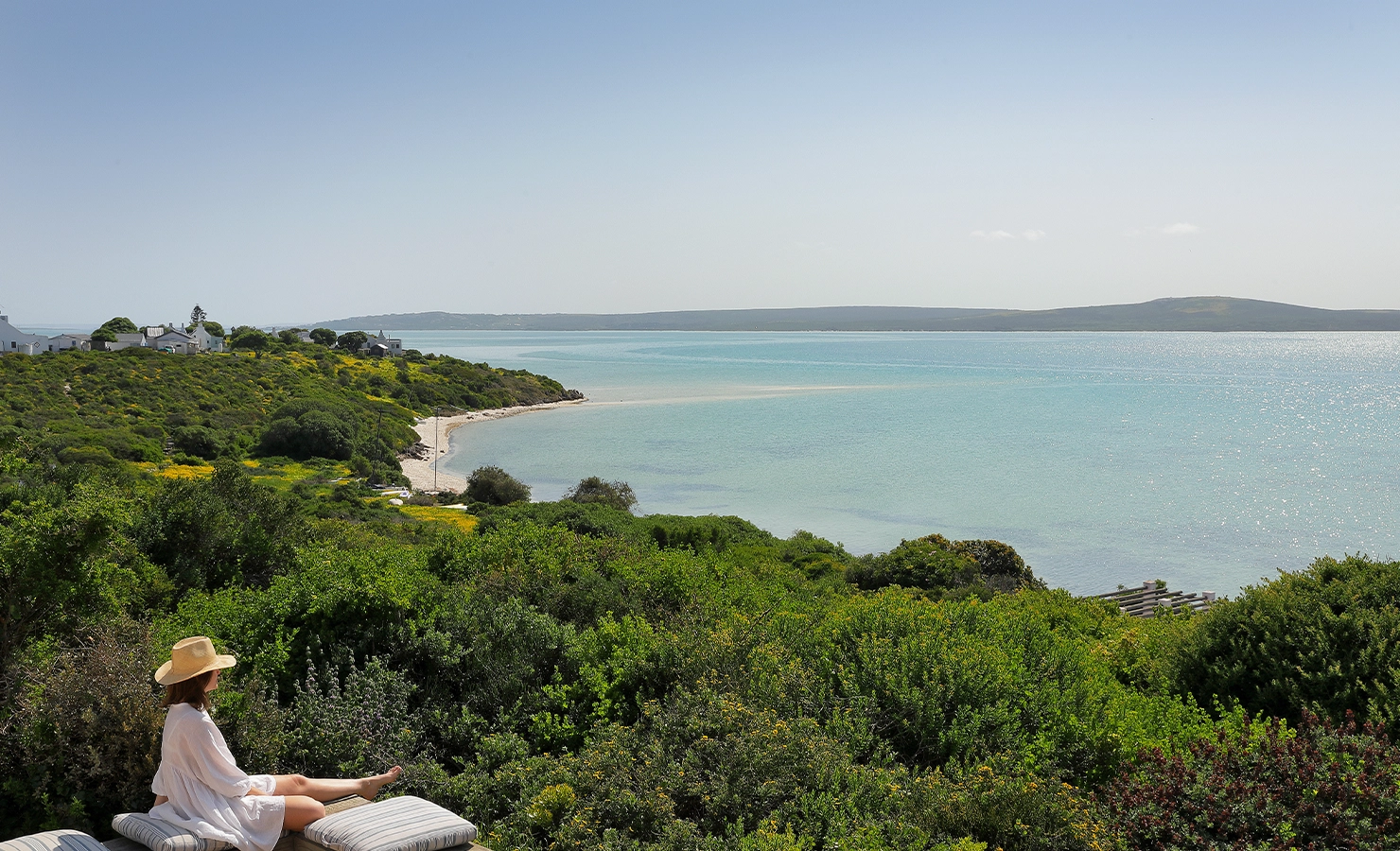Perfect Hideaways: A woman in a sunhat and white dress relaxes on a cushioned bench overlooking a serene coastal landscape with emerald fynbos, a turquoise lagoon, and whitewashed cottages nestled along the shoreline under a cloudless sky. Churchhaven, Fynbos.