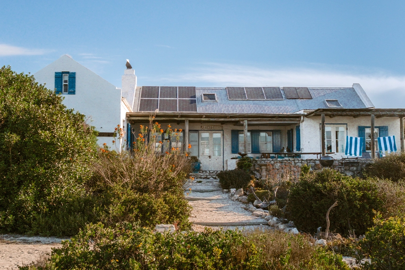 Perfect Hideaways: Whitewashed coastal cottage with blue shutters, corrugated roof fitted with solar panels and a shaded veranda above a stone terrace, reached by a sandy path through coastal fynbos. Seagull Cottage, Churchhaven.