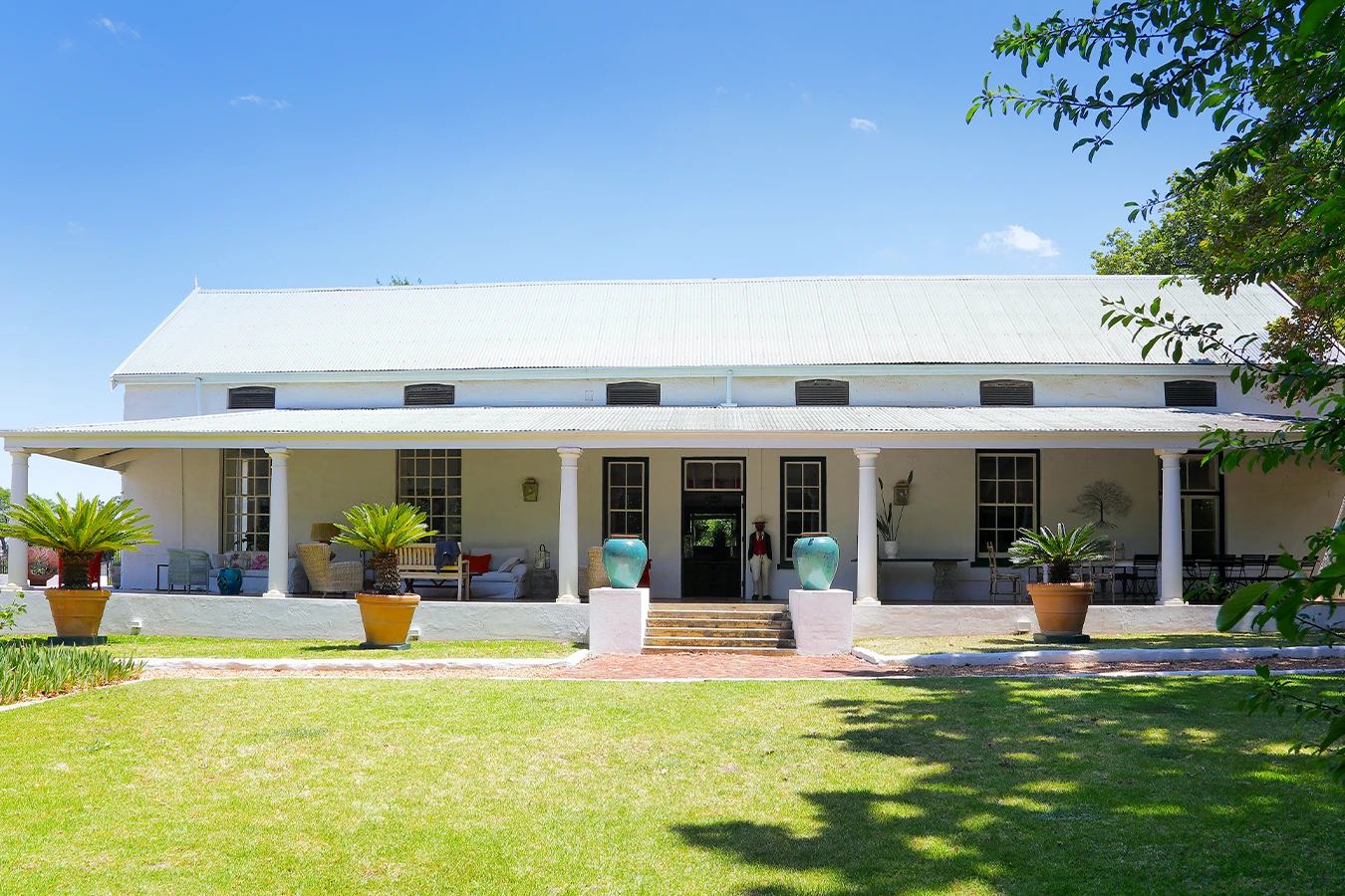 Perfect Hideaways: A charming Cape Dutch farmhouse with a wide shaded veranda, whitewashed walls, and a corrugated roof, set amidst lush green lawns and framed by potted plants and towering trees. Staart Farm, Paarl.