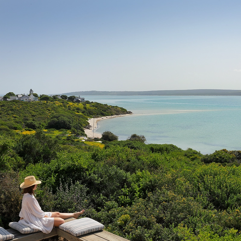 Perfect Hideaways: A woman in a sunhat and white dress relaxes on a cushioned bench overlooking a serene coastal landscape with emerald fynbos, a turquoise lagoon, and whitewashed cottages nestled along the shoreline under a cloudless sky. Churchhaven, Fynbos.