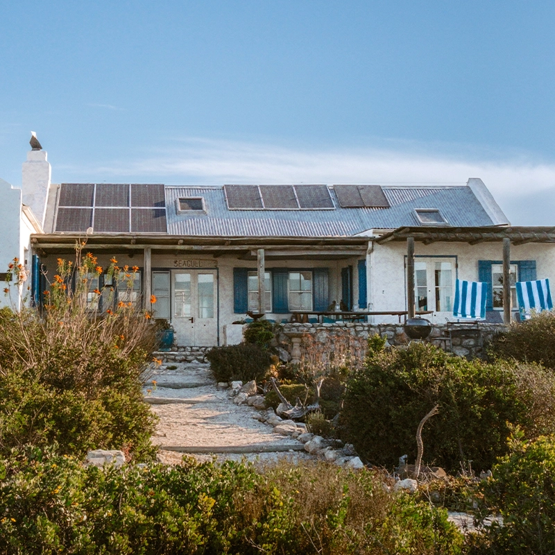 Perfect Hideaways: Whitewashed coastal cottage with blue shutters, corrugated roof fitted with solar panels and a shaded veranda above a stone terrace, reached by a sandy path through coastal fynbos. Seagull Cottage, Churchhaven.