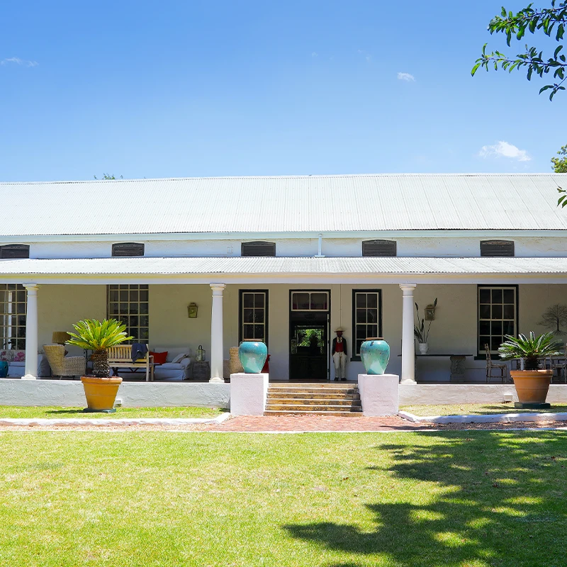 Perfect Hideaways: A charming Cape Dutch farmhouse with a wide shaded veranda, whitewashed walls, and a corrugated roof, set amidst lush green lawns and framed by potted plants and towering trees. Staart Farm, Paarl.