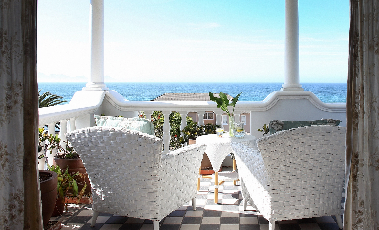Perfect Hideaways: A charming seaside balcony with white wicker chairs and a checkered floor, offering a serene view of the ocean. A small table with a jug of lemon water and fresh greenery adds to the tranquil coastal atmosphere. By The Seaside, Kalk Bay.