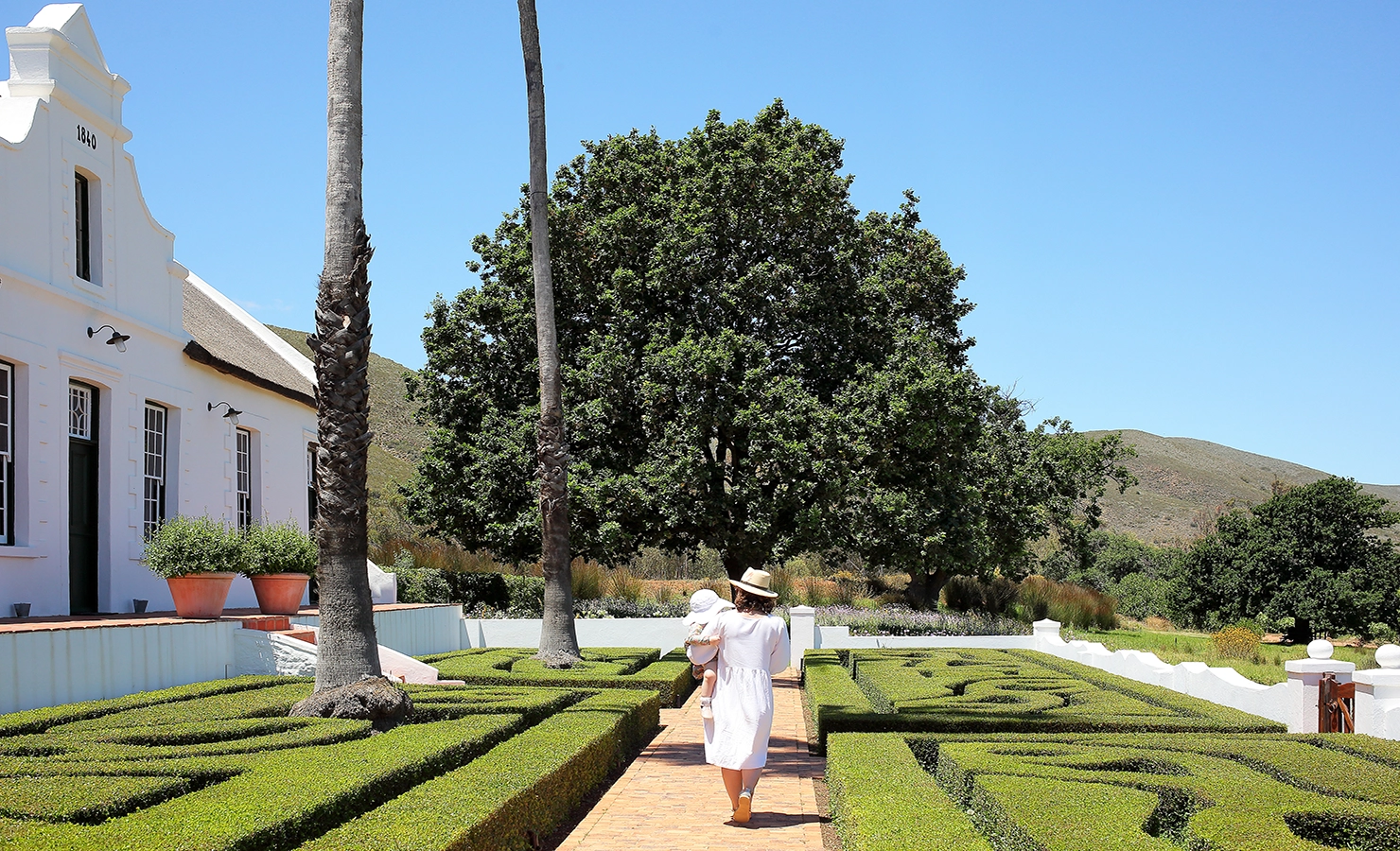 Perfect Hideaways: A woman in a white dress and sunhat walking through a manicured hedge garden with a baby, towards the main house. Penhill Manor House, Nuy Valley.