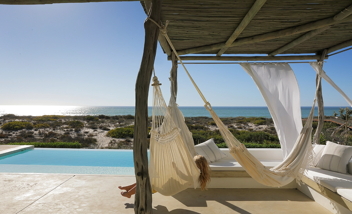 Perfect Hideaways: A serene coastal escape featuring a hammock gently swaying under a rustic wooden pergola, with views of a shimmering pool, sun-dappled dunes, and the endless Atlantic Ocean beyond – the ultimate setting for barefoot beachside relaxation. Casa Blu, Dwarskersbos.