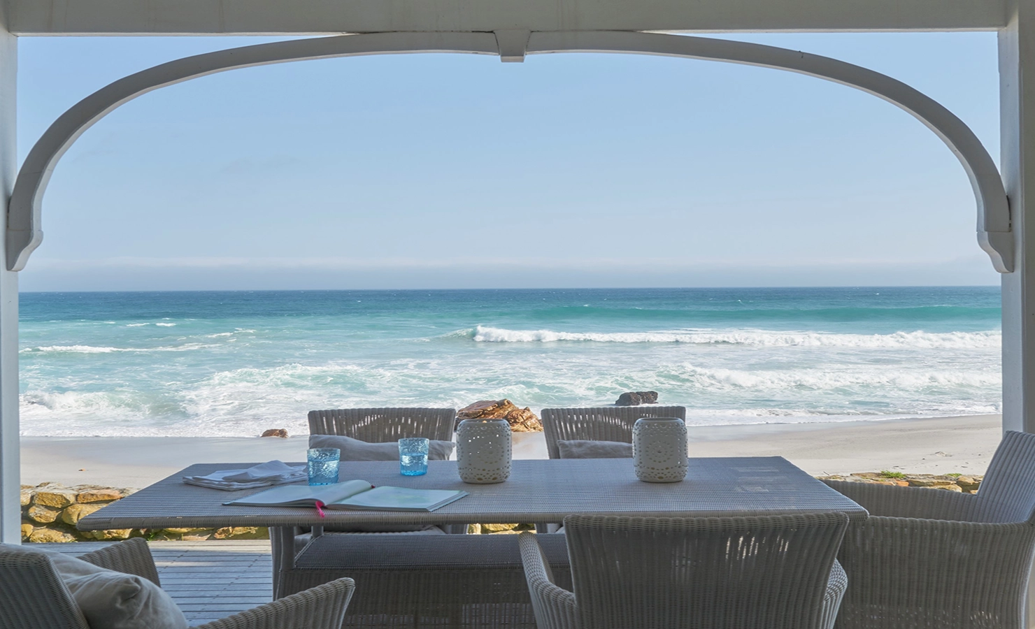 Perfect Hideaways: A serene oceanfront veranda with wicker furniture overlooks the turquoise waves and soft white sands, framed by a charming arched beam, offering the ultimate seaside escape. The Beach House, Misty Cliffs.