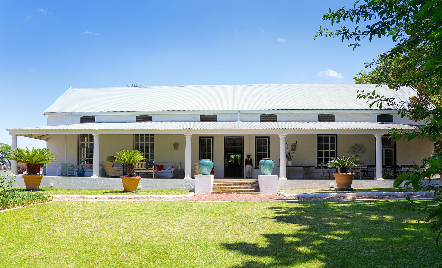 Perfect Hideaways: A charming Cape Dutch farmhouse with a wide shaded veranda, whitewashed walls, and a corrugated roof, set amidst lush green lawns and framed by potted plants and towering trees. Staart Farm, Paarl.