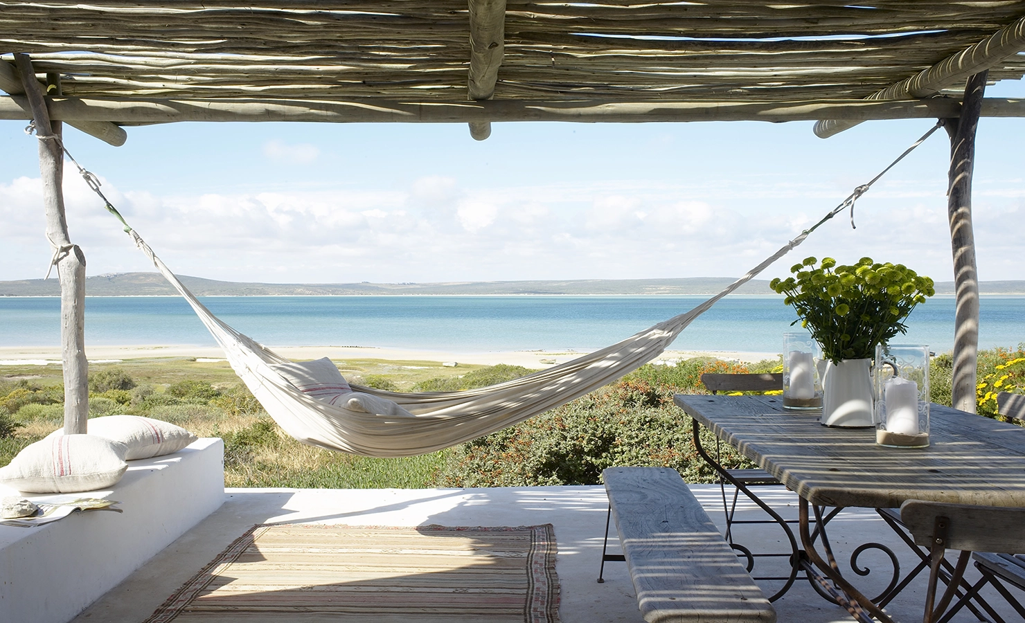 Perfect Hideaways: A serene coastal retreat featuring a shaded terrace with a rustic wooden pergola, a hammock gently swaying in the breeze, and a weathered dining table adorned with fresh flowers and candles. The view stretches out to a pristine beach and the calm, azure waters of the lagoon under a clear blue sky. Whalers Way, Churchhaven.