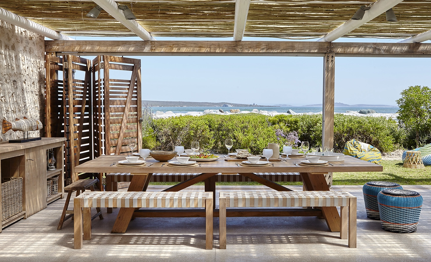 Perfect Hideaways: A beautifully styled outdoor dining area under a rustic pergola, featuring a long wooden table set for a seaside feast with woven benches, artisanal decor, and an ocean view framed by wooden shutters and lush greenery. Blue Lagoon, Langebaan.