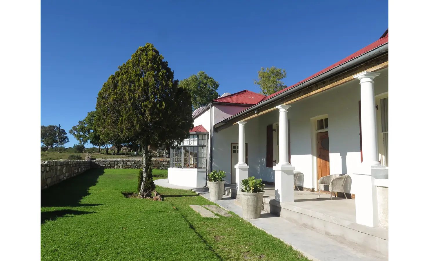 Perfect Hideaways exterior view of a white country house with red roof and pillared veranda, surrounded by manicured green lawn and stone garden walls beneath a clear blue sky. The Poplars, Sneeuberg Nature Reserve.