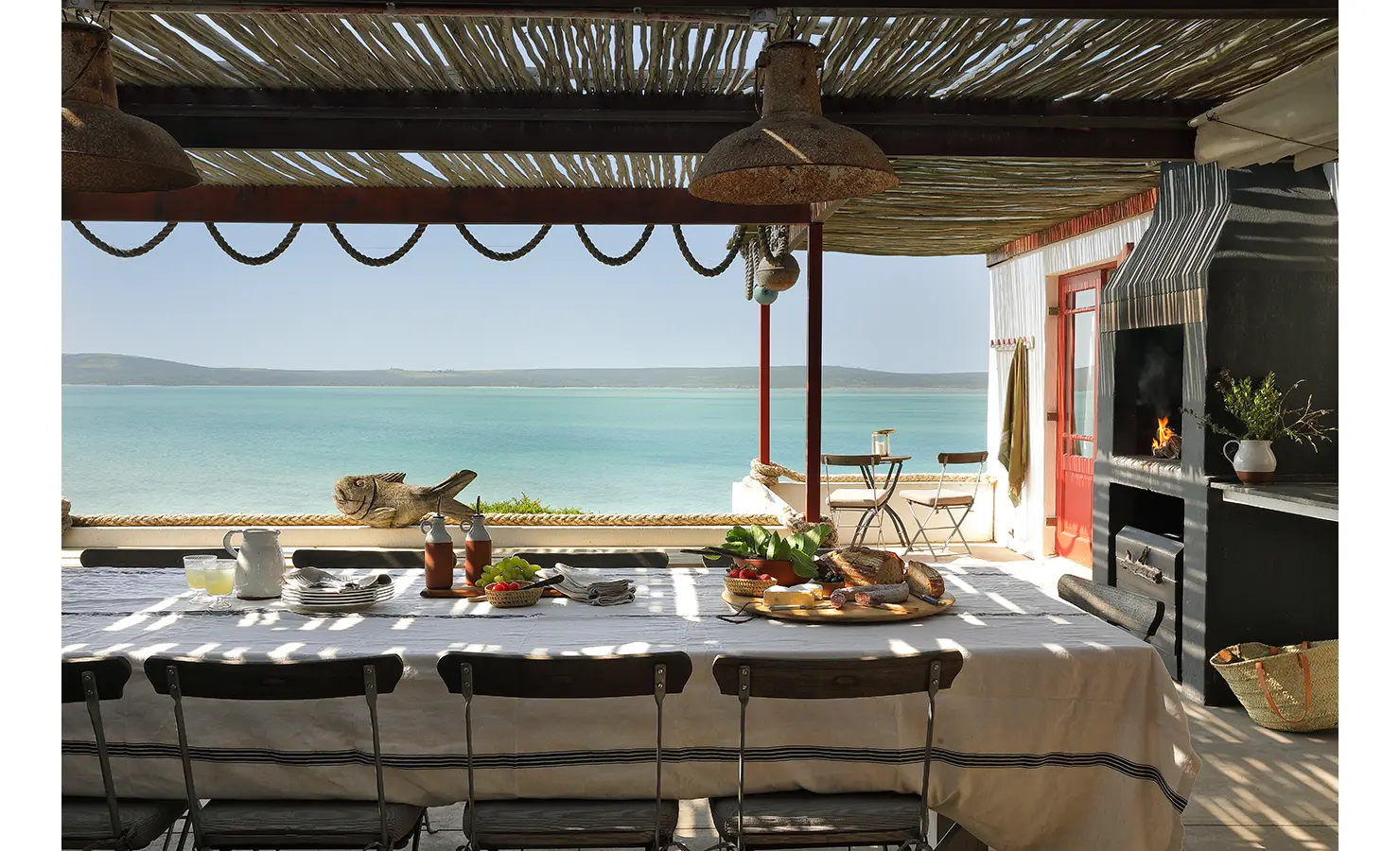 Perfect Hideaways: A long rustic dining table under a reed-covered pergola is elegantly set for a relaxed seaside meal, offering uninterrupted views of the turquoise lagoon and distant hills beyond. Churchhaven, Fynbos.