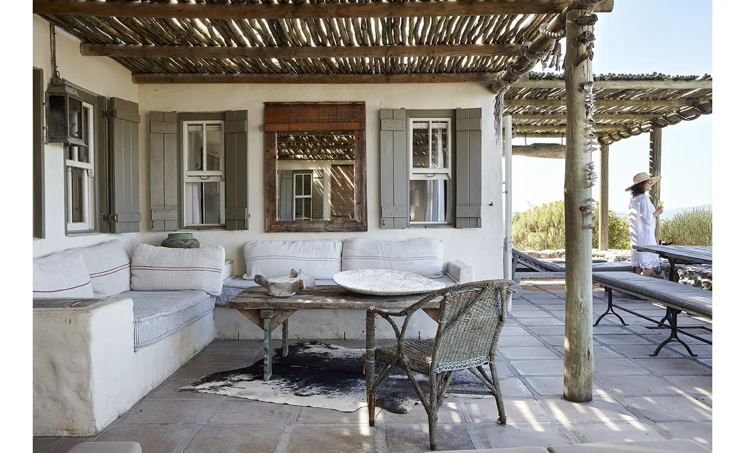 Perfect Hideaways: Shaded stoep with whitewashed built-in sofas, rustic timber table and woven chair beneath a reed pergola, grey shutters and shell garlands. Seagull Cottage, Churchhaven.