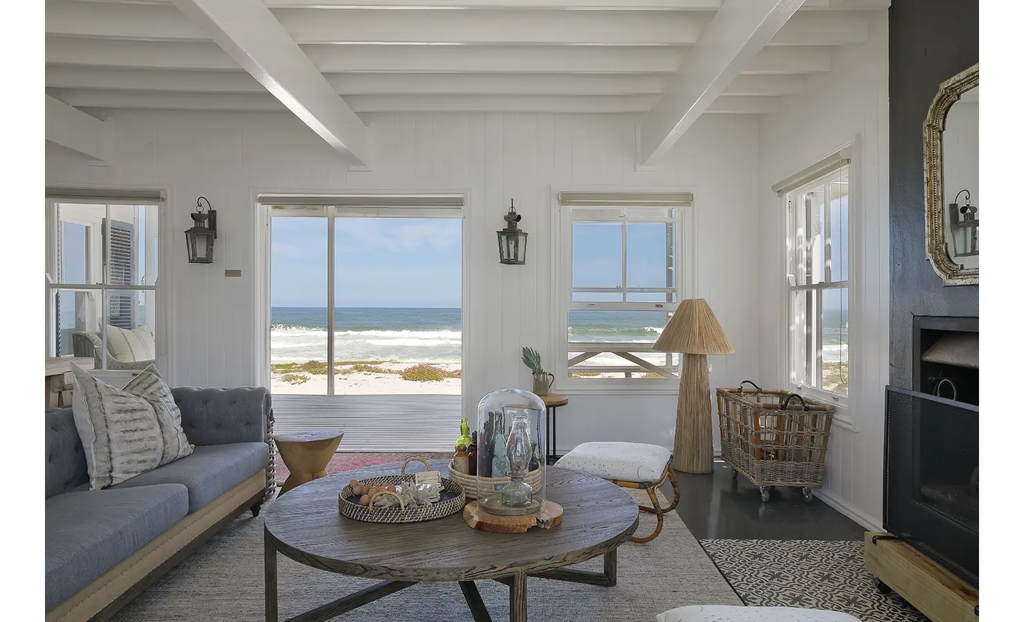 Perfect Hideaways — A serene coastal lounge featuring soft grey-blue sofas, a rustic round coffee table and sweeping views of the white sand beach through large windows. Natural textures and soft tones blend seamlessly with the wild Atlantic seascape just beyond the deck. Yzerfontein, Not Forgotten.