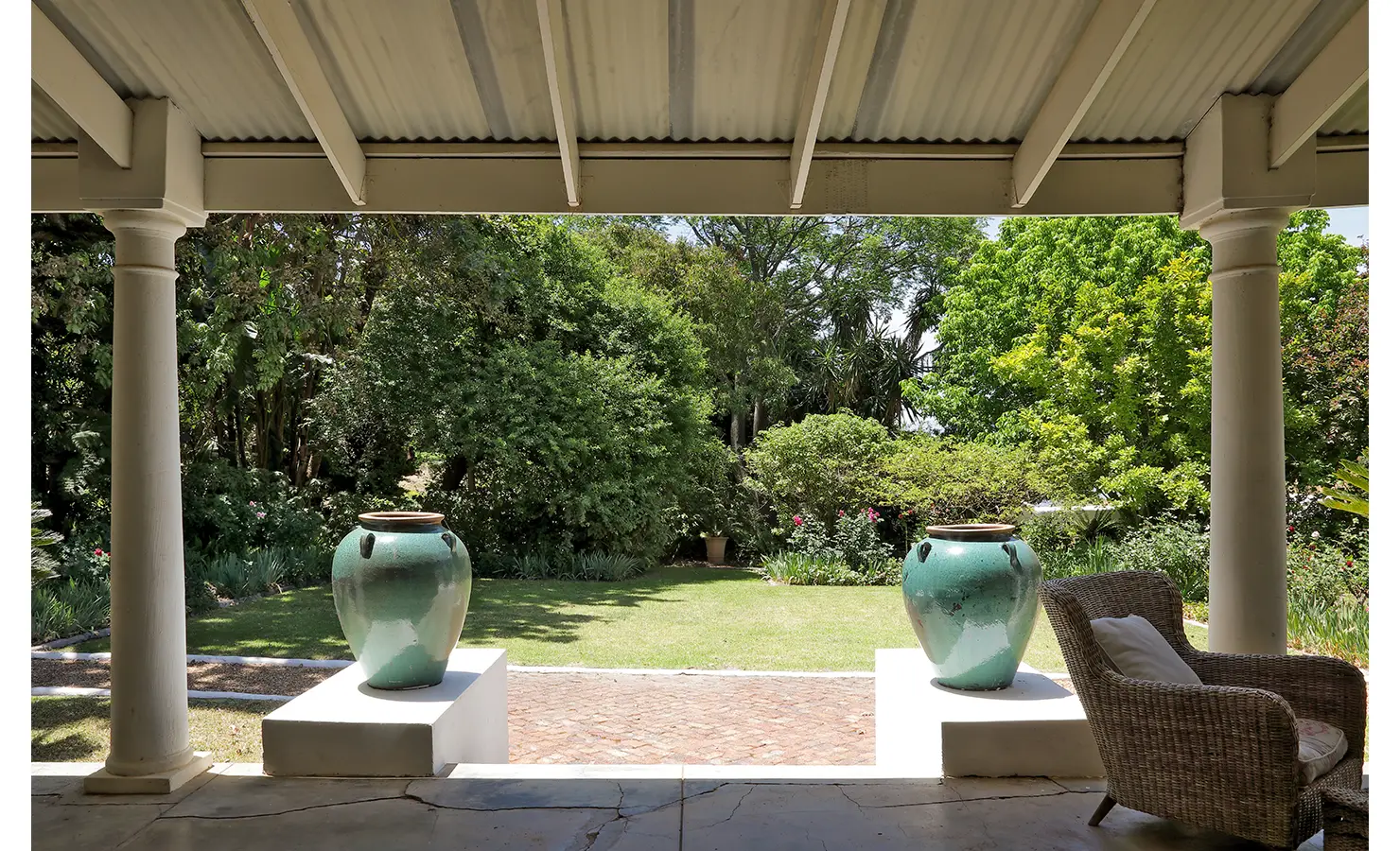 Perfect Hideaways: A shaded veranda framed by white columns overlooks a lush garden with mature trees, two striking turquoise urns, and a cosy wicker chair for relaxation. Staart Farm, Paarl.