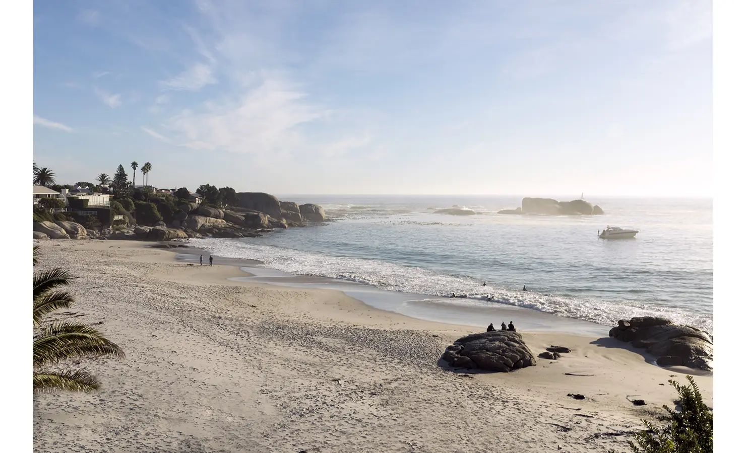 Perfect Hideaways: A pristine stretch of white sand leads to turquoise waters in this tranquil beach view, with a few walkers and a boat anchored offshore, surrounded by iconic granite boulders and palms. Clifton Bungalow 47, Clifton.