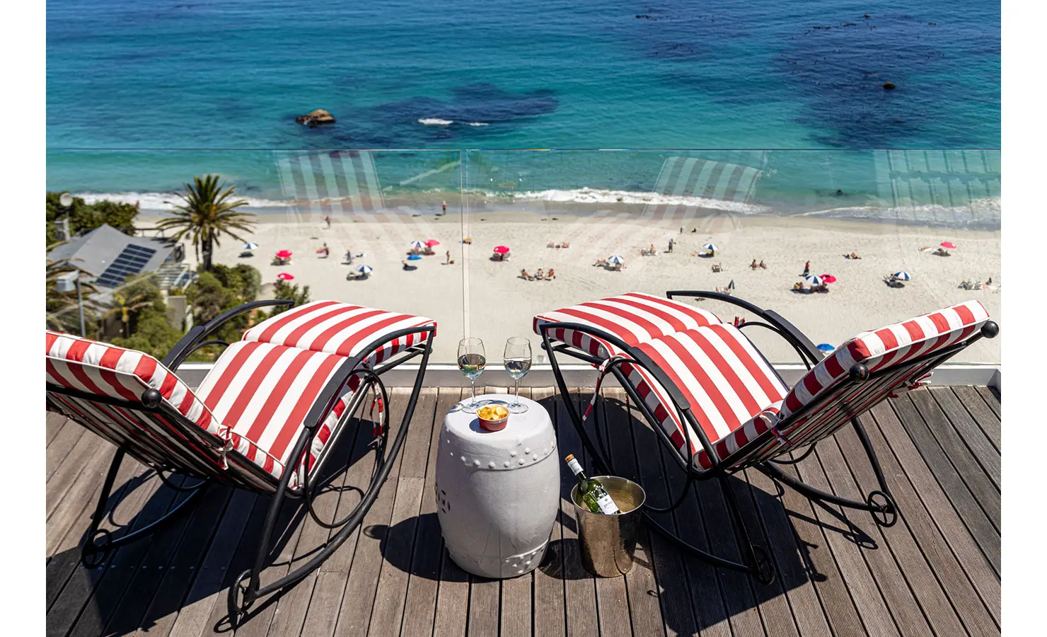 Perfect Hideaways: Two red-and-white striped sun loungers face a glass balustrade with uninterrupted views of a golden beach and the turquoise Atlantic Ocean, complete with beachgoers and parasols. A small table between the loungers holds chilled wine and snacks. Ezulwini, Clifton.