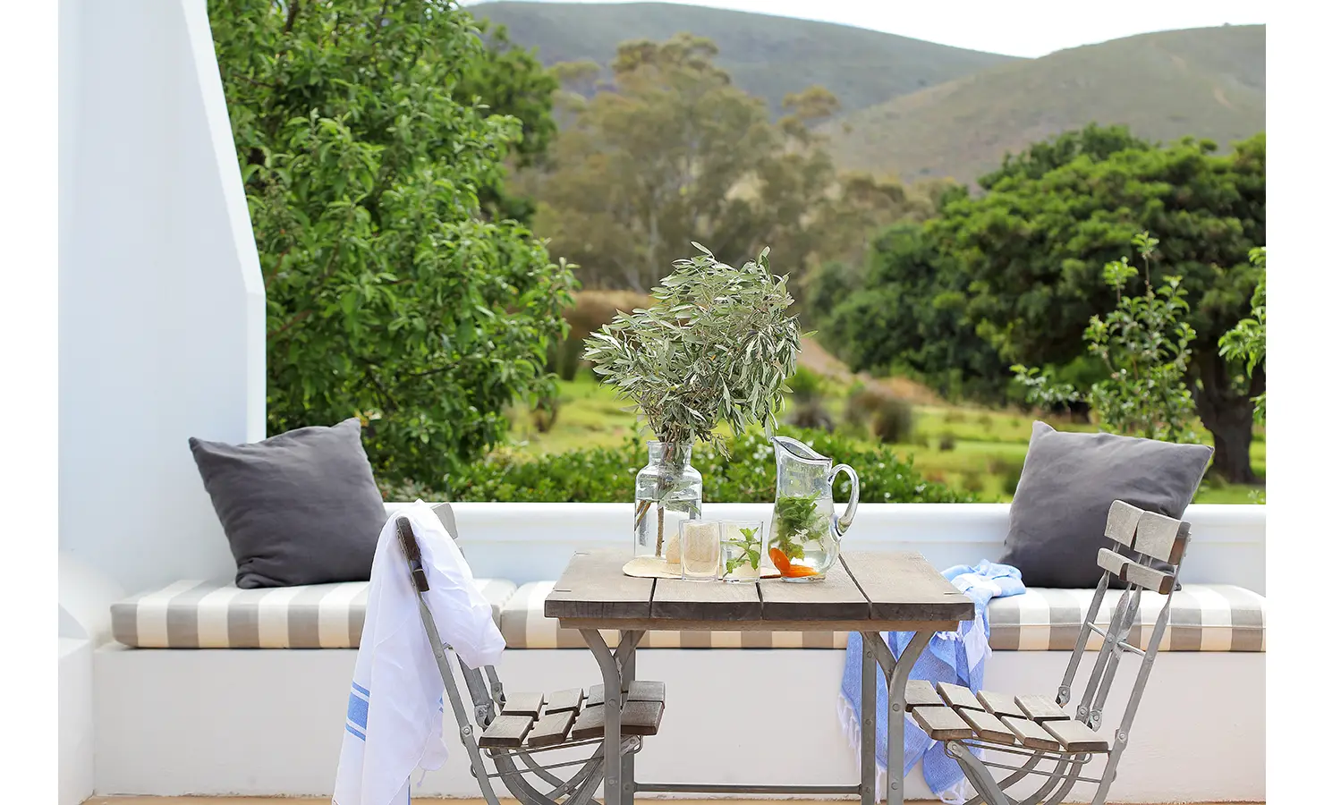 Perfect Hideaways: A charming outdoor dining nook with a rustic wooden table and wrought-iron chairs, framed by lush greenery and rolling hills. A refreshing jug of citrus-infused water and a sprig of olive branches add to the serene, countryside ambience. Penhill Fruit Barn, Nuy Valley.