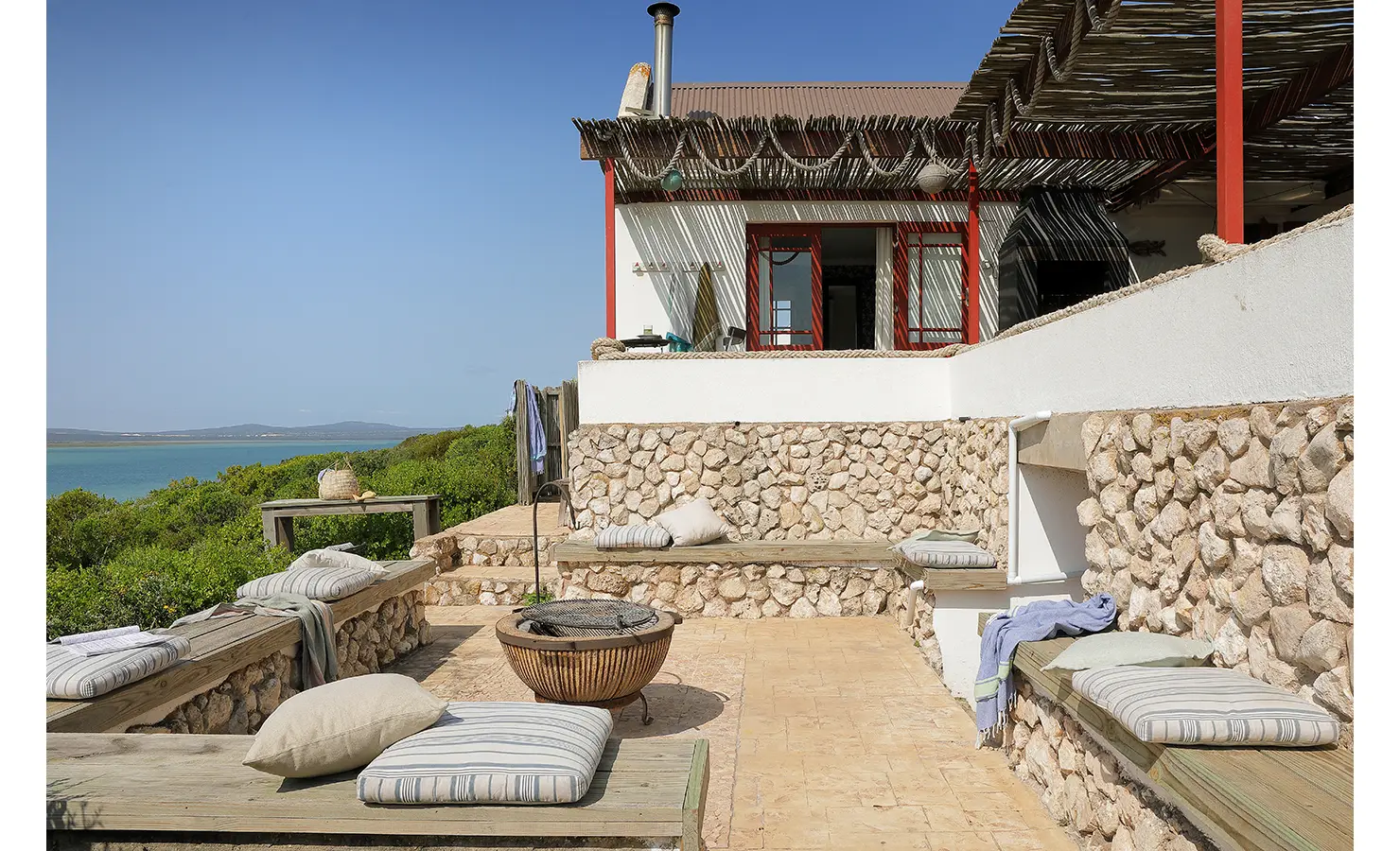 Perfect Hideaways: A stone-clad terrace with striped cushions and a central fire pit leads to a whitewashed coastal cottage with red-framed windows, nestled amid lush indigenous vegetation. Churchhaven, Fynbos.