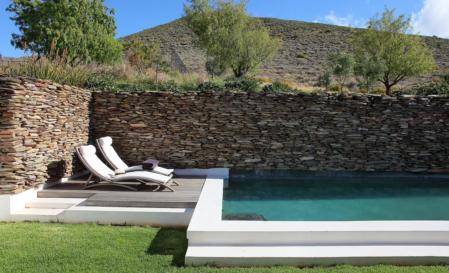 Perfect Hideaways: Two white sun loungers beside a sparkling turquoise pool, framed by a rustic stone wall and surrounded by greenery. Penhill Manor House, Nuy Valley.