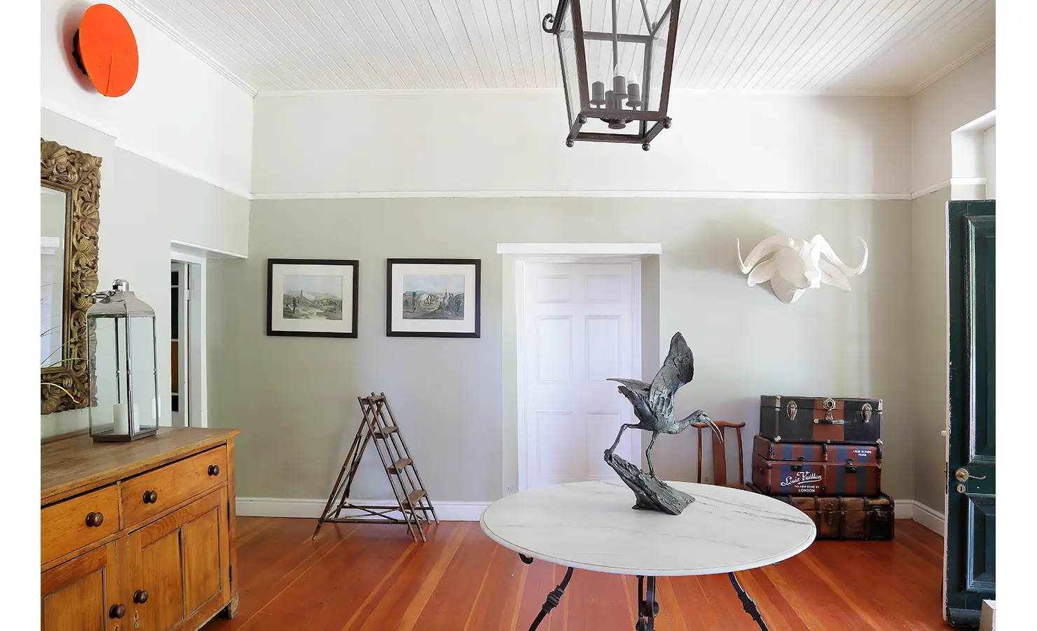 Perfect Hideaways: A charming entrance hall with wooden floors, a marble-topped round table, vintage suitcases, and eclectic décor, including a sculpted bird and framed artwork. Staart Farm, Paarl.