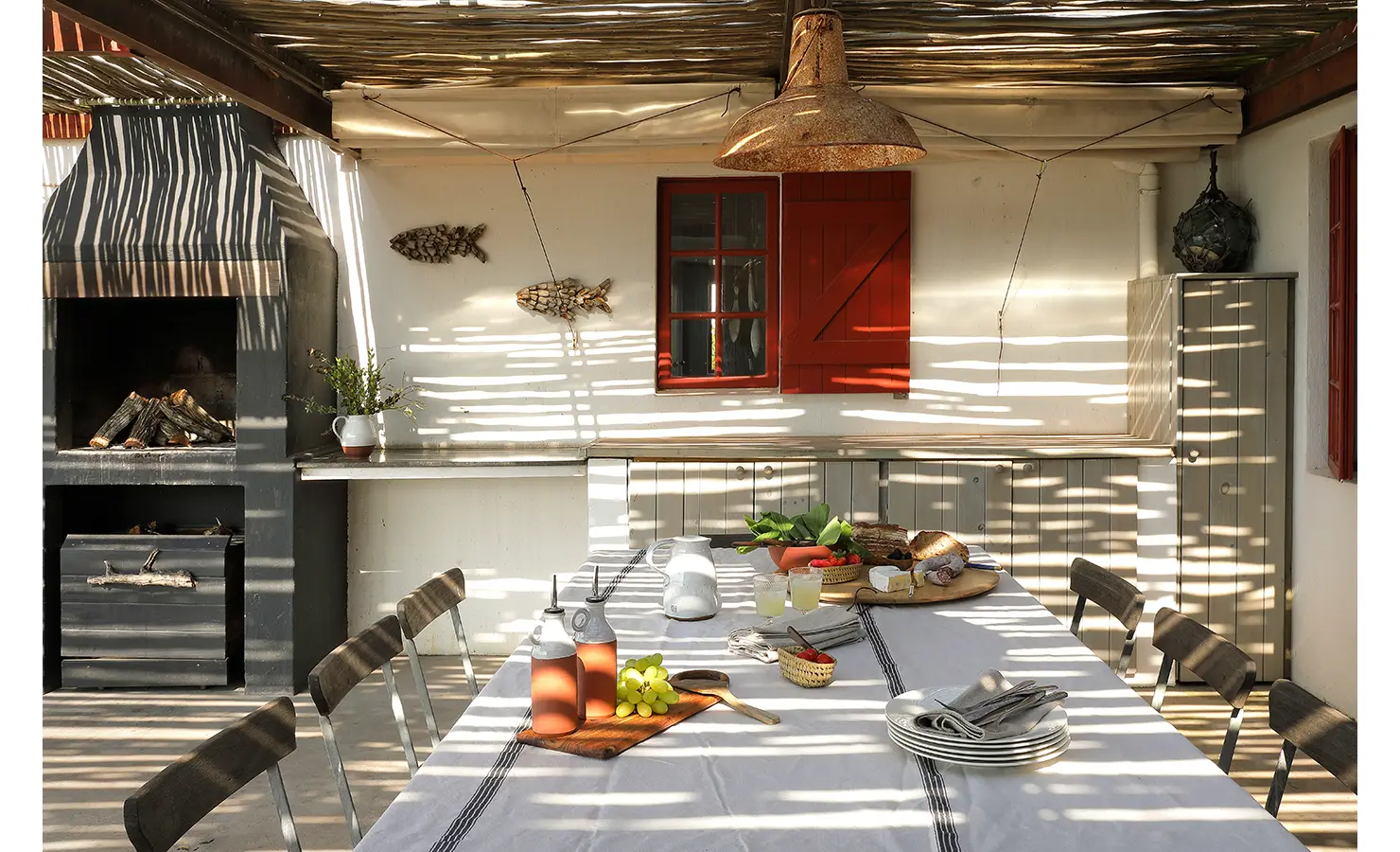 Perfect Hideaways: A shaded al fresco dining area featuring a wood-fired braai, woven chairs, and a table adorned with fresh produce and linen, bathed in dappled light filtering through a slatted canopy. Churchhaven, Fynbos.