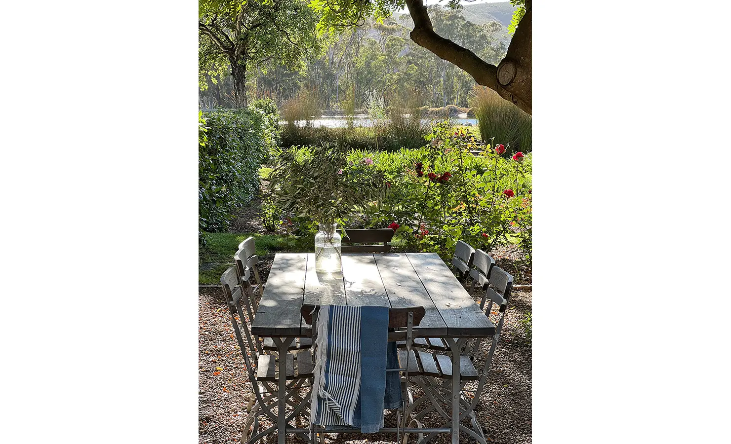 Perfect Hideaways: A charming outdoor dining setup under the shade of trees, with views of a lush garden and water feature in the distance. Penhill Manor House, Nuy Valley.