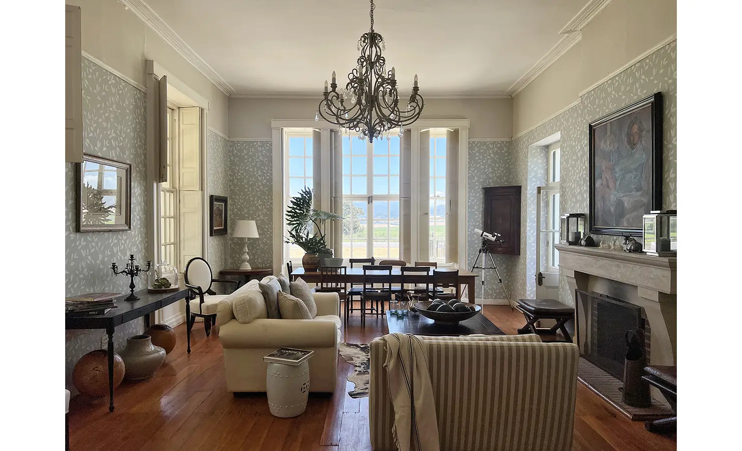 Perfect Hideaways: A grand living and dining space with high ceilings, intricate wallpaper, an elegant chandelier, and large windows that flood the room with natural light and views of the landscape. Staart Farm, Paarl.