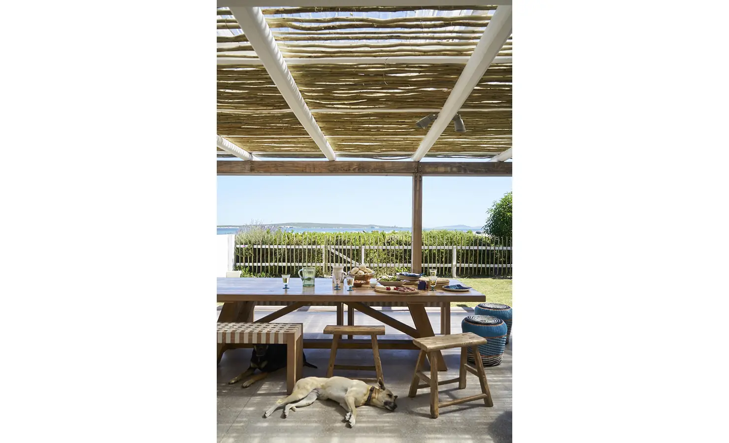 Perfect Hideaways: A shaded outdoor dining area with a wooden table set for a meal, woven benches, rustic stools, and two dogs resting beneath, all with a view of the sea. Blue Lagoon, Langebaan.
