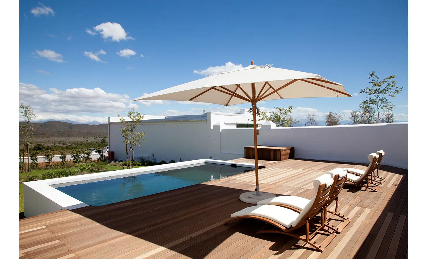 Perfect Hideaways: A stylish wooden pool deck with luxurious sun loungers and a large parasol, overlooking a breathtaking expanse of rolling hills and open sky. The crisp white walls contrast beautifully with the natural wood tones, creating an idyllic relaxation spot. Penhill Fruit Barn, Nuy Valley.