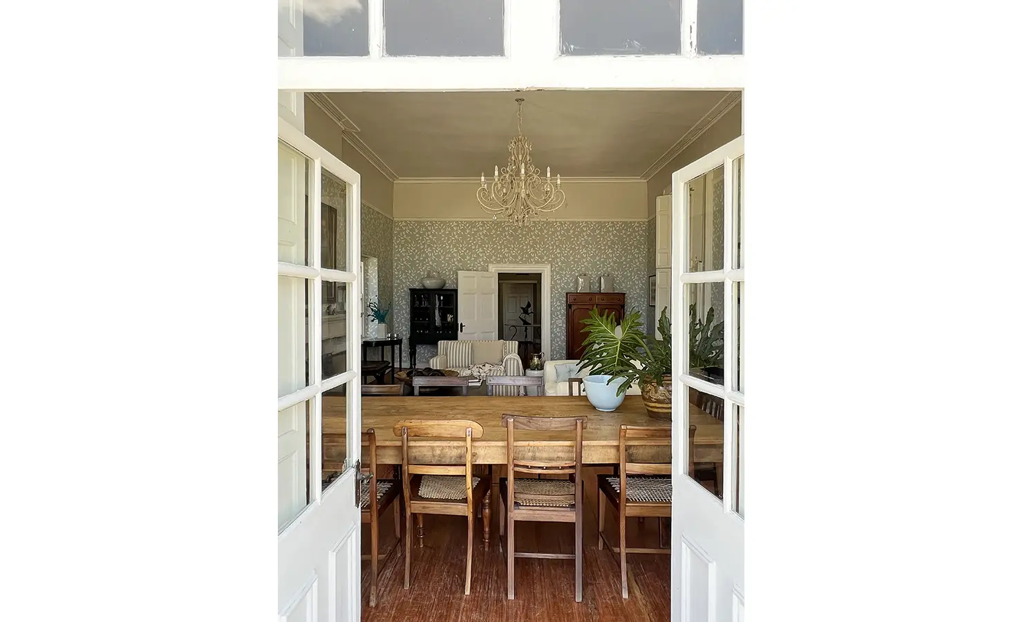 Perfect Hideaways: A dining room view through classic French doors, leading into an inviting space with a long wooden table, rustic chairs, and a chandelier casting a soft glow. Staart Farm, Paarl.