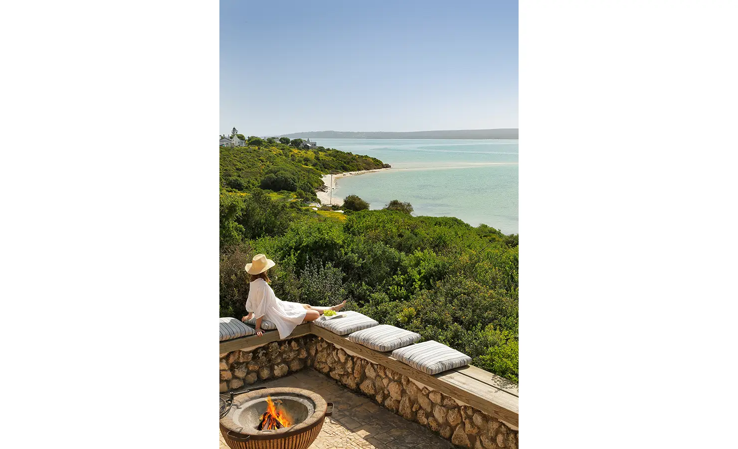 Perfect Hideaways: A woman in a flowing white dress and straw hat lounges beside a glowing fire pit, gazing across vivid green fynbos toward the curve of a quiet, sandy bay. Churchhaven, Fynbos.