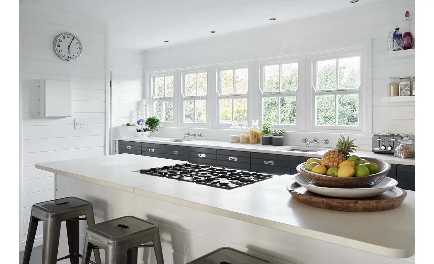 Perfect Hideaways: A bright, contemporary kitchen with a large central island and gas hob, surrounded by windows and finished with crisp white counters, dark drawers, and industrial bar stools. Noordhoek Beach House, Noordhoek.