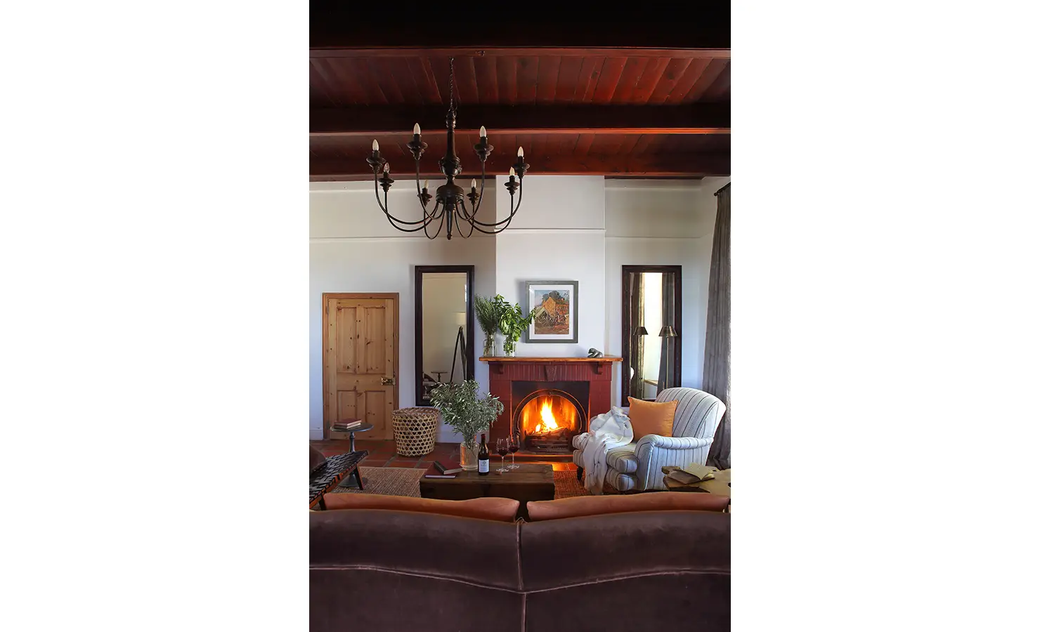 Perfect Hideaways: A cosy living room with a roaring fireplace, plush seating, and elegant decor, highlighted by a chandelier. Penhill Manor House, Nuy Valley.