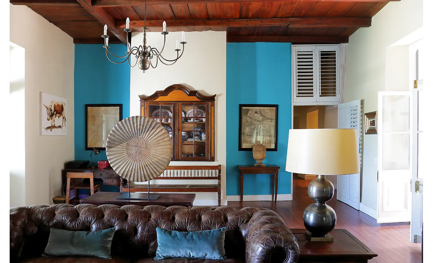 Perfect Hideaways: A cosy yet refined lounge with a deep-buttoned leather couch, antique furniture, a wooden ceiling, and a striking turquoise accent wall adding a modern touch. Staart Farm, Paarl.