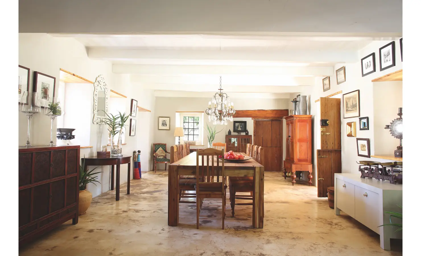 Spacious dining room with a solid wooden dining table, smooth epoxy flooring and decorated with trinkets and wall art with a statement chandelier.