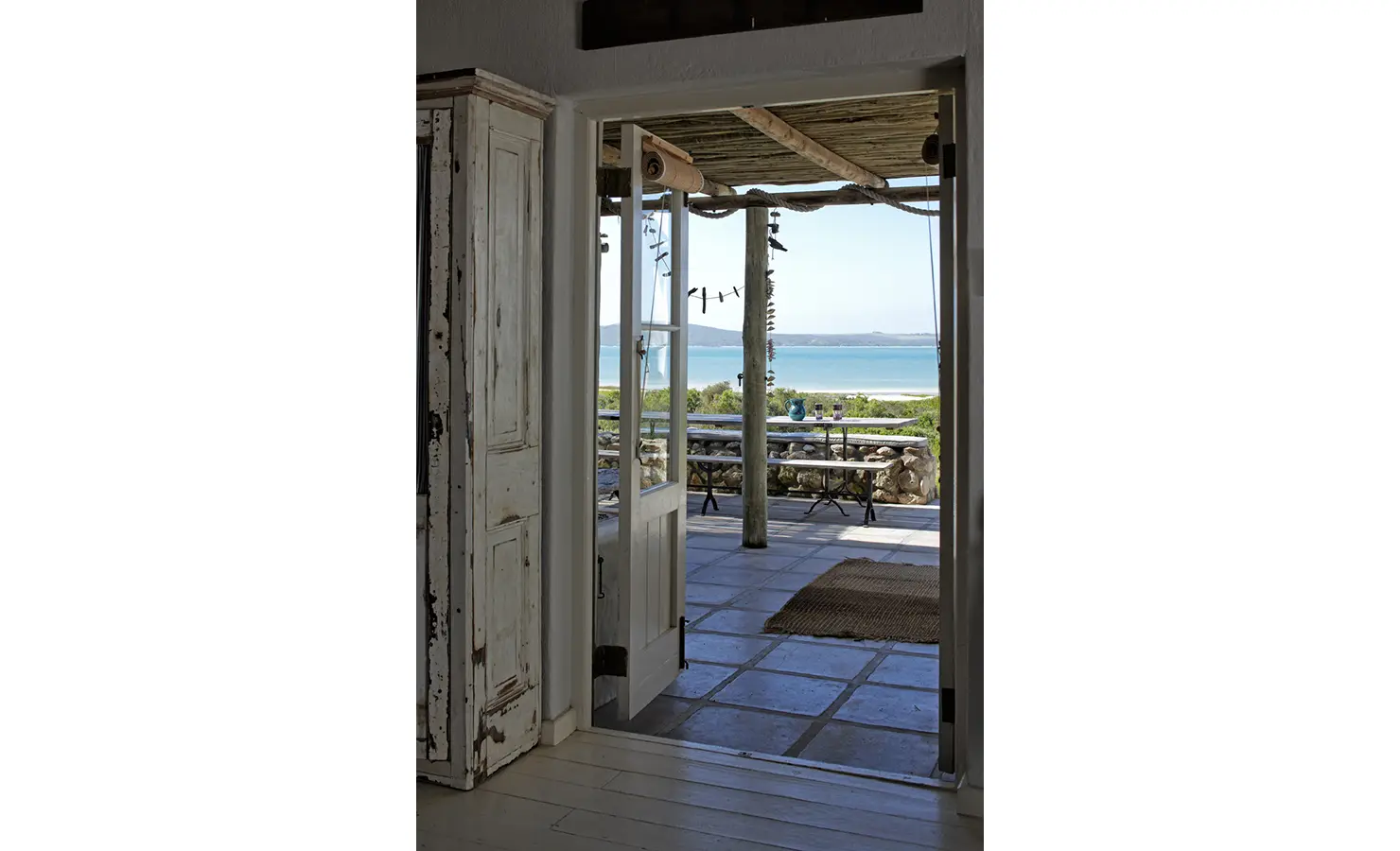 Perfect Hideaways: A view through open double doors revealing a covered patio with seating, leading out to a peaceful beachside setting. Seagull Cottage, Churchhaven.