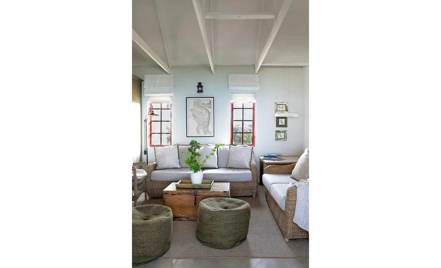 Perfect Hideaways: A sun-drenched sitting room with natural textures and botanical accents features pale woven sofas, wooden chests, and light streaming in through red-framed windows. Churchhaven, Fynbos.