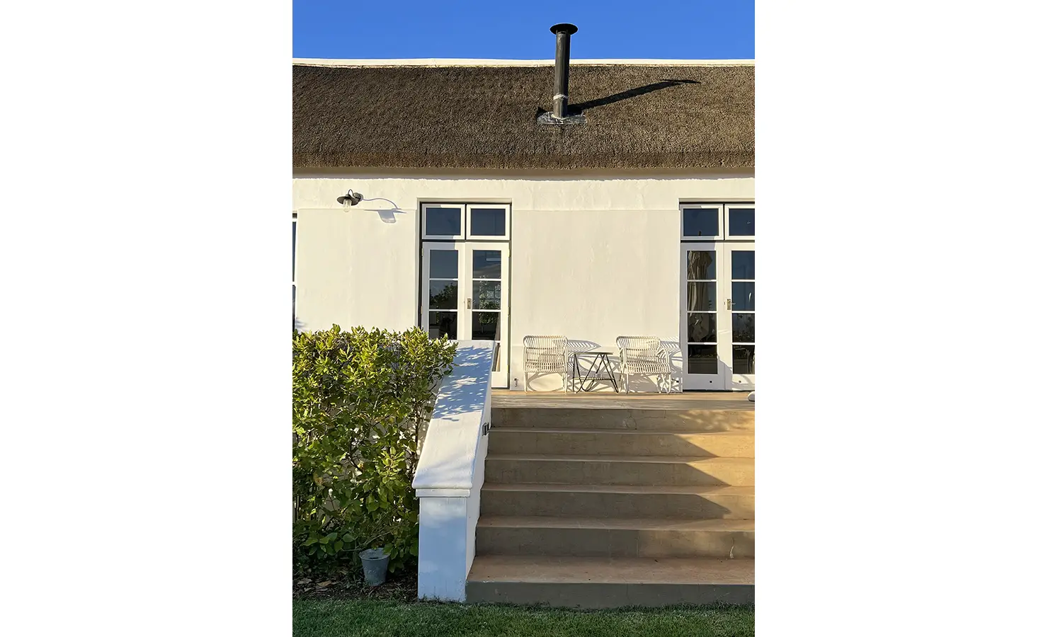 Perfect Hideaways: A sunlit veranda with white French doors leading into a Cape Dutch-style home. The inviting outdoor seating area offers a perfect spot to enjoy a morning coffee while overlooking the manicured garden. Penhill Fruit Barn, Nuy Valley.