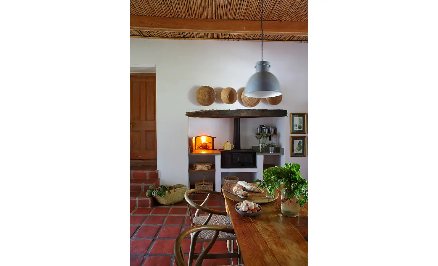 Perfect Hideaways: A rustic kitchen with a wood-burning oven, woven baskets, and a wooden dining table adorned with fresh produce. Penhill Manor House, Nuy Valley.