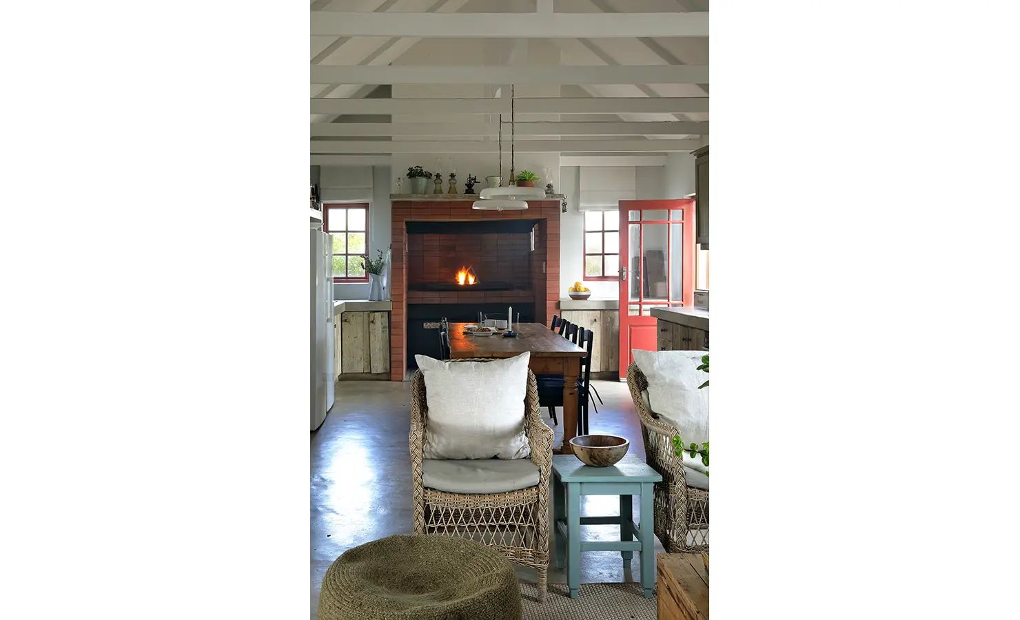 Perfect Hideaways: A welcoming open-plan kitchen and dining space with exposed beams, vintage cabinetry, a central fireplace, and coral-red accents infuses coastal charm with farmhouse warmth. Churchhaven, Fynbos.