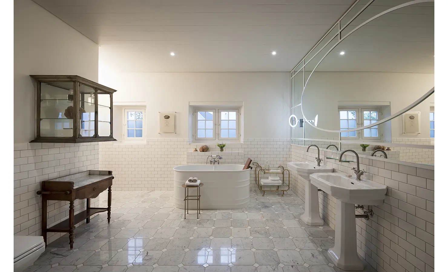 Perfect Hideaways: A spacious bathroom with marble flooring, a freestanding bathtub, classic twin pedestal sinks, and a large, uniquely shaped mirror enhancing the elegant aesthetic. Babylonstoren, Franschhoek.