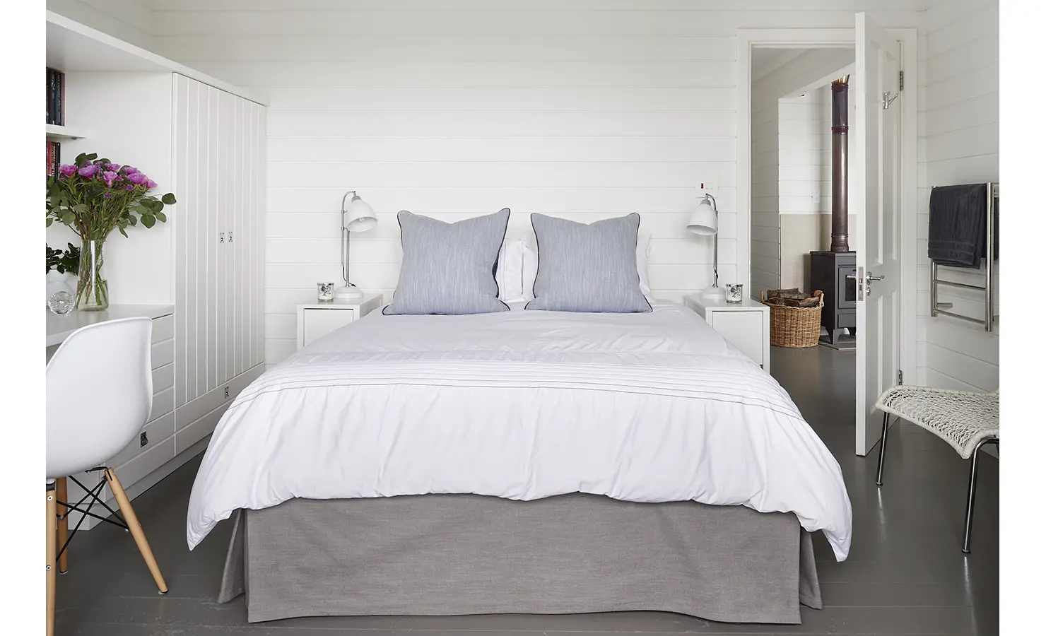 Perfect Hideaways: A guest bedroom with clean lines and soft neutral tones, featuring a white and grey colour palette, built-in cabinetry, and wood-burning stove visible through the adjoining bathroom. Noordhoek Beach House, Noordhoek.
