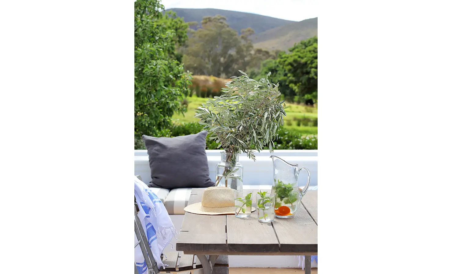 Perfect Hideaways: A beautifully styled alfresco dining setup with a rustic wooden table, cushioned seating, and a refreshing pitcher of herb-infused water. The surrounding greenery and distant mountains complete the serene countryside setting. Penhill Fruit Barn, Nuy Valley.