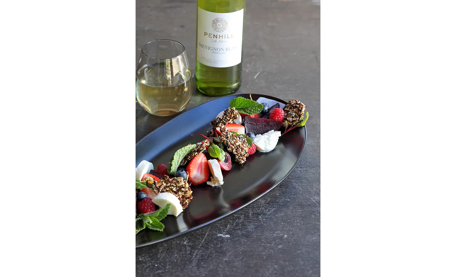 Perfect Hideaways: A beautifully plated fruit and cheese dish, accompanied by a glass and bottle of Penhill Sauvignon Blanc on a dark surface. Penhill Manor House, Nuy Valley.