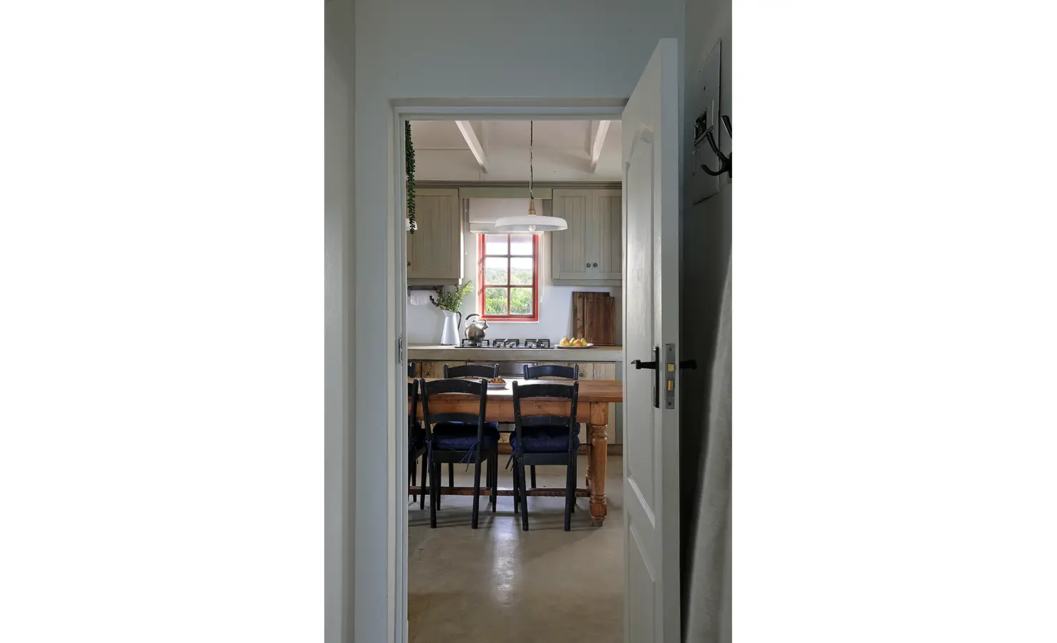 Perfect Hideaways: A peek through a white doorway reveals a country-style kitchen and dining nook, bathed in soft light from a red-framed window above rustic timber cabinetry. Churchhaven, Fynbos.