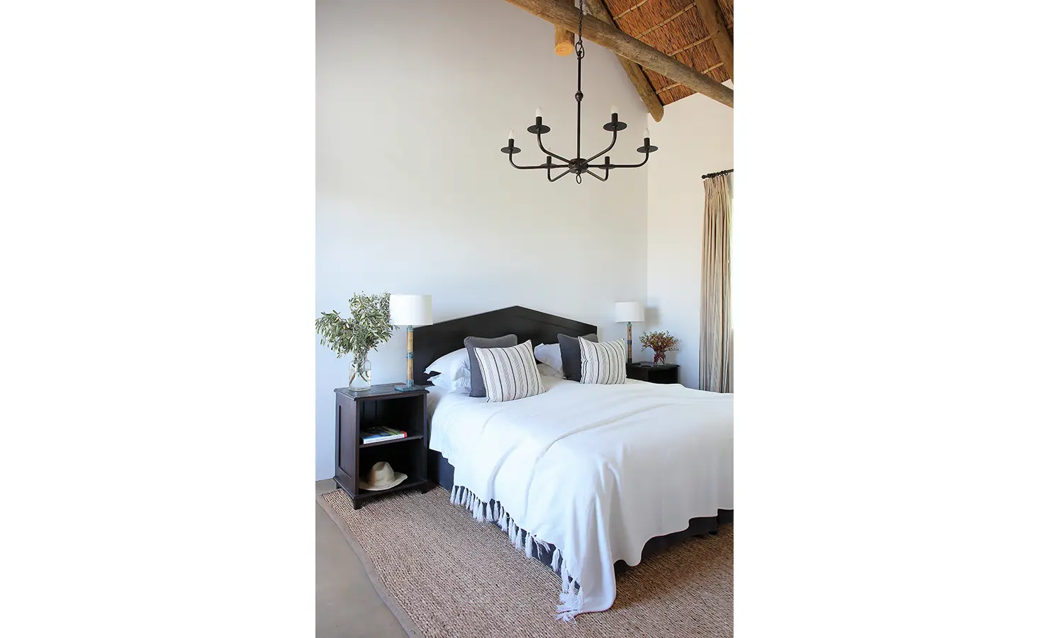 Perfect Hideaways: A bright and elegant bedroom with a high thatched ceiling, a wrought-iron chandelier, and a dark wood bed dressed in crisp white linens. Natural textures and neutral tones create a peaceful and inviting retreat. Penhill Fruit Barn, Nuy Valley.
