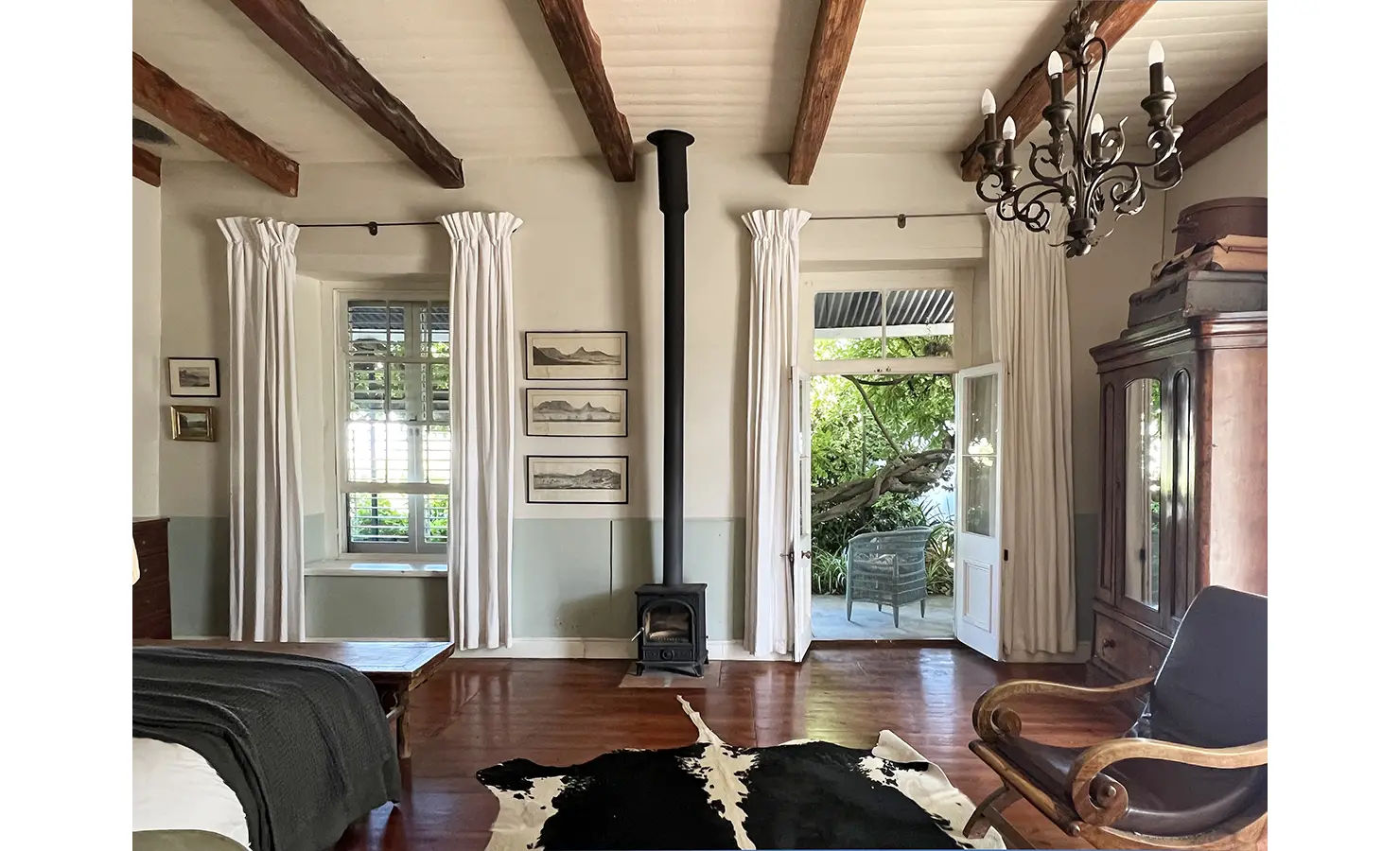 Perfect Hideaways: A tranquil bedroom with exposed wooden beams, a vintage chandelier, a wood-burning stove, and large French doors opening onto a shaded garden terrace. Staart Farm, Paarl.