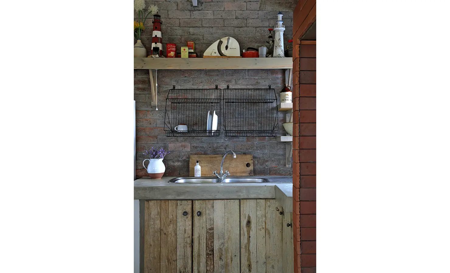 Perfect Hideaways: A compact scullery corner with a double sink, exposed brick walls, open shelves, and nautical ornaments captures the laid-back, eclectic spirit of coastal living. Churchhaven, Fynbos.