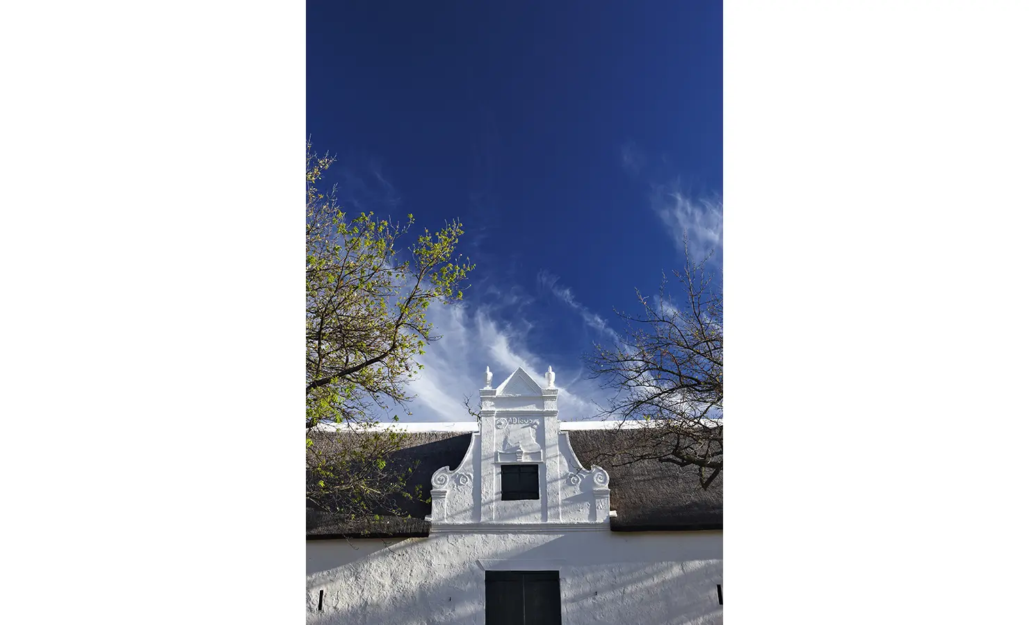 Perfect Hideaways: The gabled façade of a historic Cape Dutch homestead, painted in crisp white, set against a deep blue sky with wispy clouds and framed by leafy trees. Babylonstoren, Franschhoek.