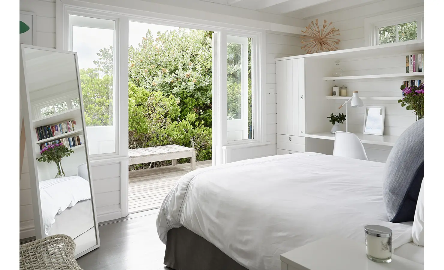 Perfect Hideaways: A tranquil bedroom opening onto a wooden deck framed by indigenous foliage, with white built-ins, a large mirror, and soft blue and white accents. Noordhoek Beach House, Noordhoek.